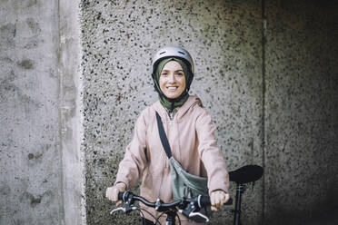 Porträt einer lächelnden jungen Frau mit Fahrrad, die in der Nähe einer Mauer steht - MASF33224