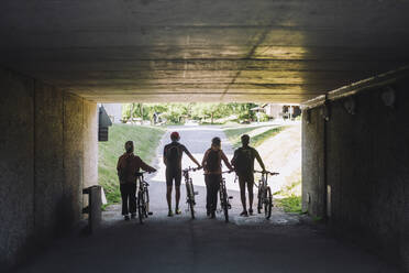Male and female friends wheeling with cycles while walking through underpass - MASF33225