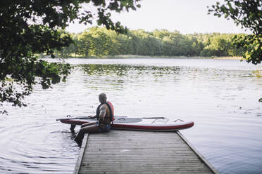 Mann mit Paddelboard schaut auf den See, während er auf dem Steg sitzt - MASF33228