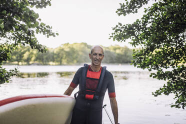 Älterer Mann mit Neoprenanzug und Schwimmweste, der ein Paddleboard hält - MASF33232