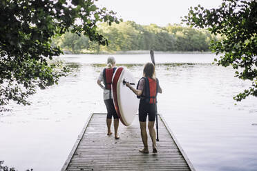 Ältere Frauen tragen ein Paddelbrett, während sie auf dem Steg zum See gehen - MASF33236
