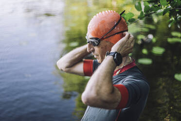 Männlicher Sportler bereitet sich auf das Schwimmen vor, während er seine Badekappe im See anpasst - MASF33240