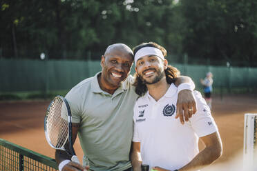 Glücklicher Mann mit Arm um männlichen Freund mit Stirnband auf dem Tennisplatz - MASF33256