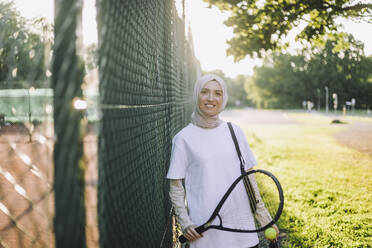 Porträt einer lächelnden Frau, die einen Tennisschläger am Zaun hält - MASF33259