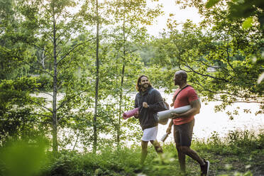 Männliche Freunde tragen Turnmatten und unterhalten sich beim Spaziergang im Wald - MASF33271