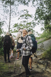 Seitenansicht einer lächelnden älteren Frau, die beim Wandern mit Freunden im Wald einen Stock hält - MASF33274