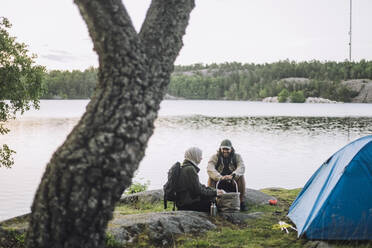 Friends talking with each other while sitting on rock by tent at lakeshore - MASF33277