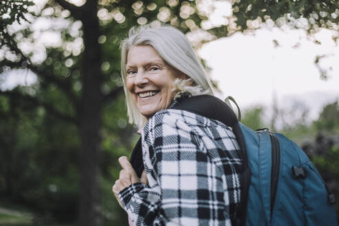 Portrait of happy senior woman looking over shoulder carrying backpack while hiking - MASF33284