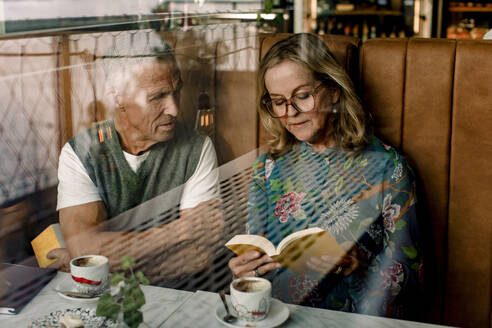 Ältere Frau liest einem männlichen Freund ein Buch vor, der mit verschränkten Armen in einem Café sitzt, gesehen durch Glas - MASF33328
