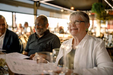 Lächelnde ältere Frau mit Brille sitzt mit Menükarte bei Männern im Restaurant - MASF33329