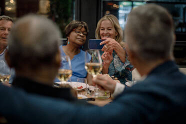Lächelnde ältere Frauen fotografieren männliche Freunde mit Weingläsern in einem Restaurant - MASF33337