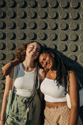 Smiling teenage girl with arm around female friend standing against wall - MASF33356