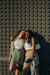 Portrait of happy teenage girl standing with arm around friend against wall during sunny day - MASF33357