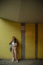 Full length of smiling teenage girl standing with hands in pockets against yellow wall - MASF33361