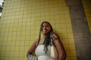 Low angle view of happy teenage girl with braided hair standing against yellow wall - MASF33362