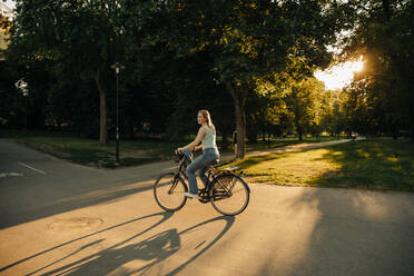 Side view of teenage girl riding bicycle on road at park during sunset - MASF33371