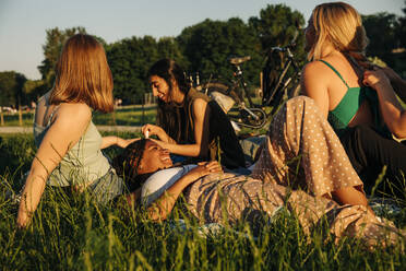 Lächelndes Teenager-Mädchen liegt im Gras und unterhält sich mit Freundinnen im Park bei Sonnenuntergang - MASF33380
