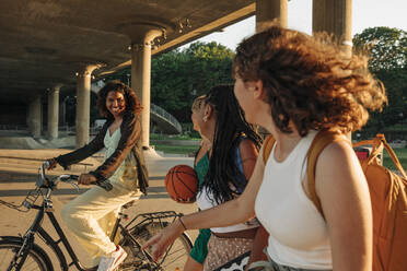 Teenager-Mädchen, die eine lächelnde Freundin betrachten, die unter einer Brücke Fahrrad fährt - MASF33389