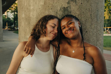 Portrait of smiling teenage girl with arm around friend standing against column - MASF33398