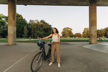Teenage girl with bicycle standing in front of bridge at park - MASF33412
