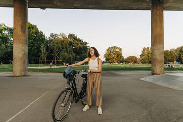 Teenager-Mädchen mit Fahrrad vor einer Brücke im Park stehend - MASF33412