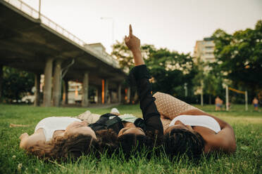 Teenager-Mädchen mit erhobener Hand, die auf einen Punkt zeigt, während sie inmitten von Freunden auf dem Rasen eines Parks liegt - MASF33419