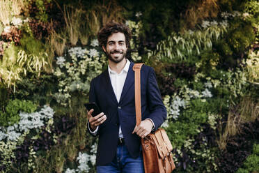 Smiling businessman with shoulder bag standing in front of plants - EBBF07025