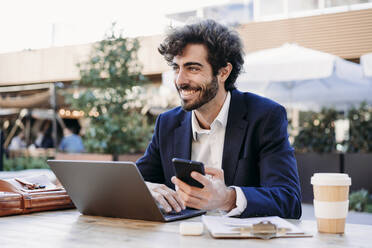 Lächelnder Geschäftsmann mit Smartphone und Laptop bei der Arbeit in einem Café - EBBF07031