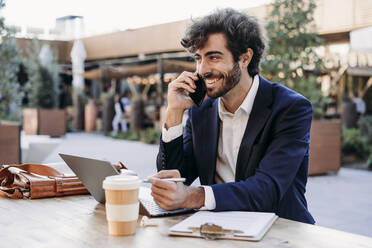 Glücklicher Geschäftsmann, der am Tisch im Café mit seinem Mobiltelefon spricht - EBBF07039