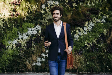 Smiling businessman with smart phone walking in front of plants - EBBF07057