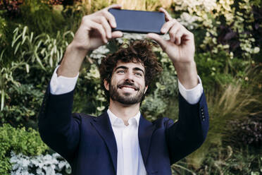 Lächelnder Geschäftsmann nimmt Selfie durch Smartphone in der Nähe von Pflanzen - EBBF07066