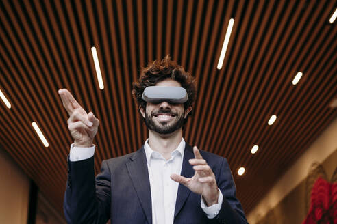 Lächelnder Geschäftsmann, der ein Virtual-Reality-Headset trägt und gestikuliert - EBBF07072