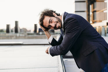 Smiling businessman with head in hand leaning on railing - EBBF07083