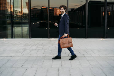Businessman with briefcase walking on footpath by modern building - EBBF07085