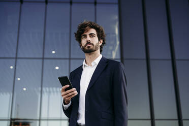 Contemplative businessman with smart phone in front of modern building - EBBF07086