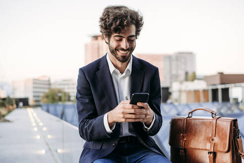 Businessman using smart phone sitting by briefcase on railing - EBBF07091