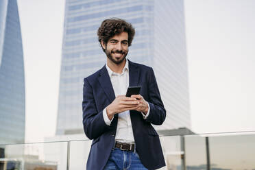 Smiling urban businessman with smart phone in front of building - EBBF07093