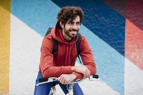 Lächelnder junger Mann sitzt auf einem Fahrrad vor einer bunten Wand - EBBF07095