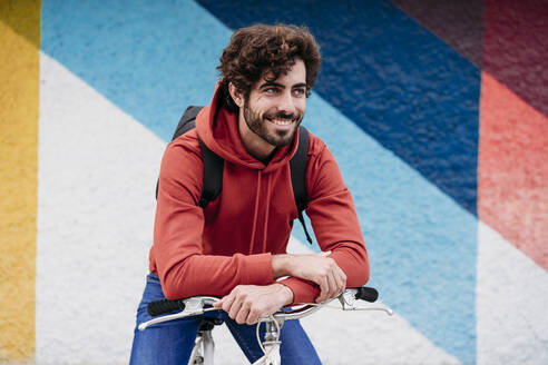 Smiling young man sitting on bicycle in front of colorful wall - EBBF07095