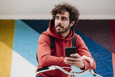 Gut aussehender Mann mit Smartphone, der sich vor einer bunten Wand auf ein Fahrrad stützt - EBBF07099