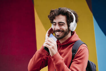 Glücklicher Mann mit geschlossenen Augen, der Musik hört und sein Smartphone vor einer bunten Wand hält - EBBF07105