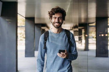 Happy man with smart phone standing in corridor - EBBF07108
