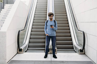 Junger Mann mit Smartphone vor einer Rolltreppe stehend - EBBF07114
