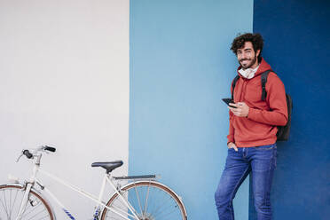 Lächelnder Mann, der sein Smartphone an einem Fahrrad an der Wand hält - EBBF07119
