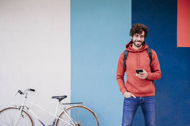 Lächelnder Mann mit Hand in der Tasche am Fahrrad vor einer bunten Wand - EBBF07121