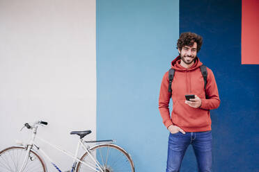 Smiling man with hand in pocket by bicycle in front of colorful wall - EBBF07121