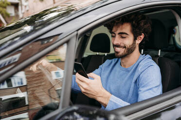 Smiling man using smart phone sitting in car - EBBF07126
