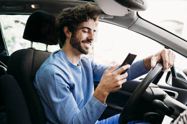 Smiling man using smart phone sitting on driver's seat in car - EBBF07127