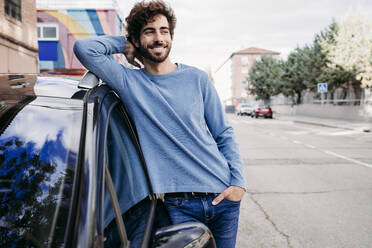 Smiling man with hand in pocket leaning on car - EBBF07133