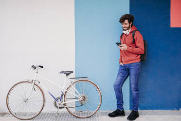 Lächelnder Mann mit Smartphone auf dem Fahrrad in der Nähe einer bunten Wand - EBBF07145